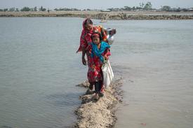 A woman and a young girl in colorful clothing walking along a narrow path surrounded by water in Bangladesh. The woman is carrying a metal pot on her shoulder.