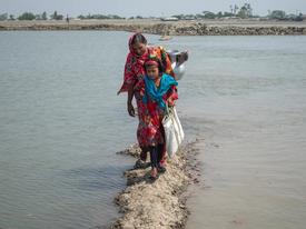 A woman and a young girl in colorful clothing walking along a narrow path surrounded by water in Bangladesh. The woman is carrying a metal pot on her shoulder.