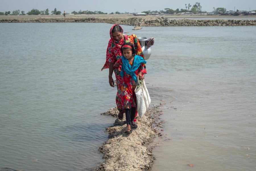 A woman and a young girl in colorful clothing walking along a narrow path surrounded by water in Bangladesh. The woman is carrying a metal pot on her shoulder.
