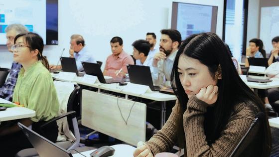 A group of people sitting at desks with laptops in a training session, focusing on their screens in a classroom setting.