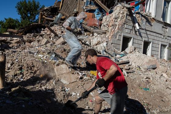 Two people working amidst rubble and debris from a destroyed building in Ukraine. One person is wearing a protective suit, and the other is shoveling debris, highlighting the aftermath of destruction.