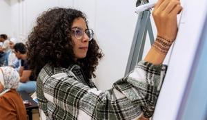 Image of a person in glasses and a plaid shirt writing on a large pad of paper in a room full of people sitting at tables.