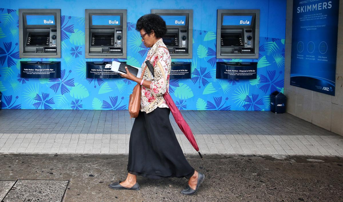 A woman walking in front of a row of ATMs on a bright blue wall with floral patterns, carrying a purse, an umbrella, and looking at her phone.