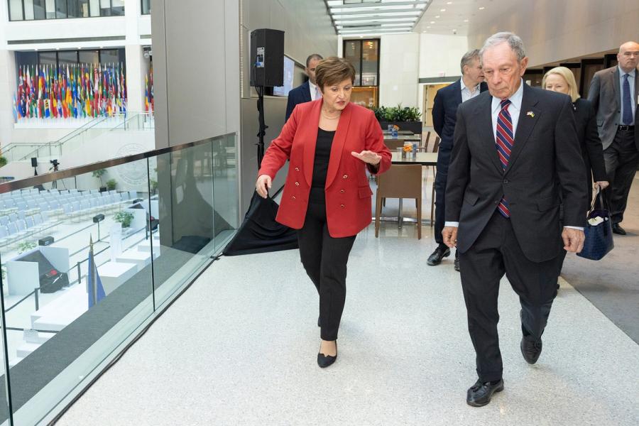 A woman in a red jacket and a man in a suit walking together in a building, with a display of international flags in the background.