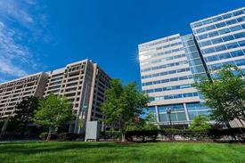 A view of two modern office buildings with large glass windows under a bright blue sky, surrounded by green trees and a well-maintained grassy area.