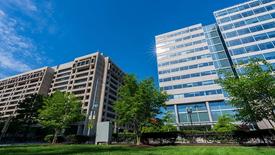 A view of two modern office buildings with large glass windows under a bright blue sky, surrounded by green trees and a well-maintained grassy area.