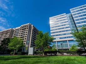 A view of two modern office buildings with large glass windows under a bright blue sky, surrounded by green trees and a well-maintained grassy area.