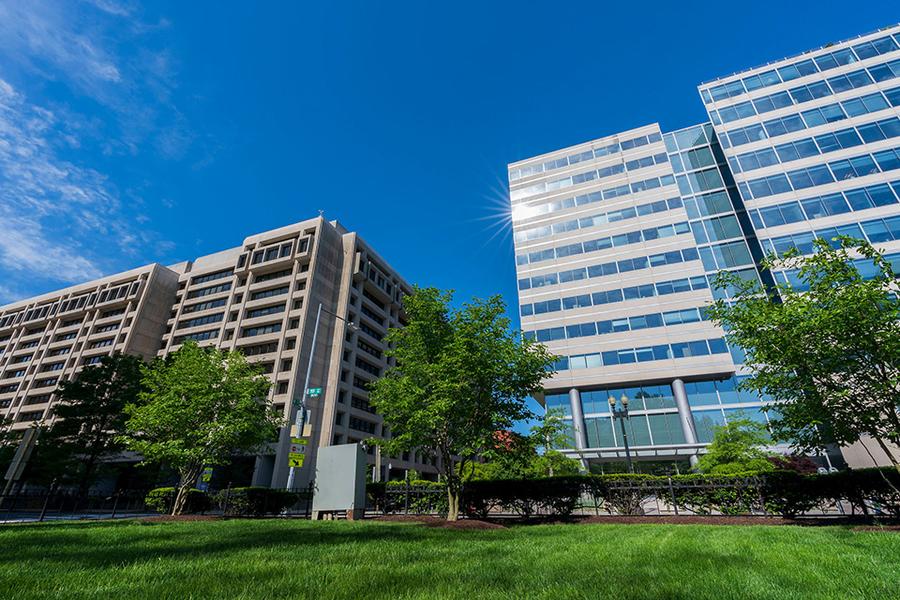 A view of two modern office buildings with large glass windows under a bright blue sky, surrounded by green trees and a well-maintained grassy area.