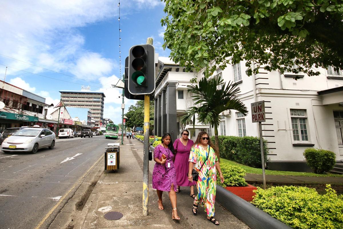 Three people walking on a sidewalk in a city in Fiji, with a traffic light, cars on the road, and buildings in the background on a sunny day.