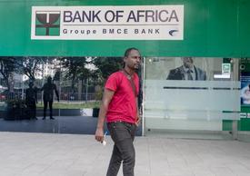A man walking in front of a Bank of Africa branch in Ghana, wearing a red shirt and holding papers.