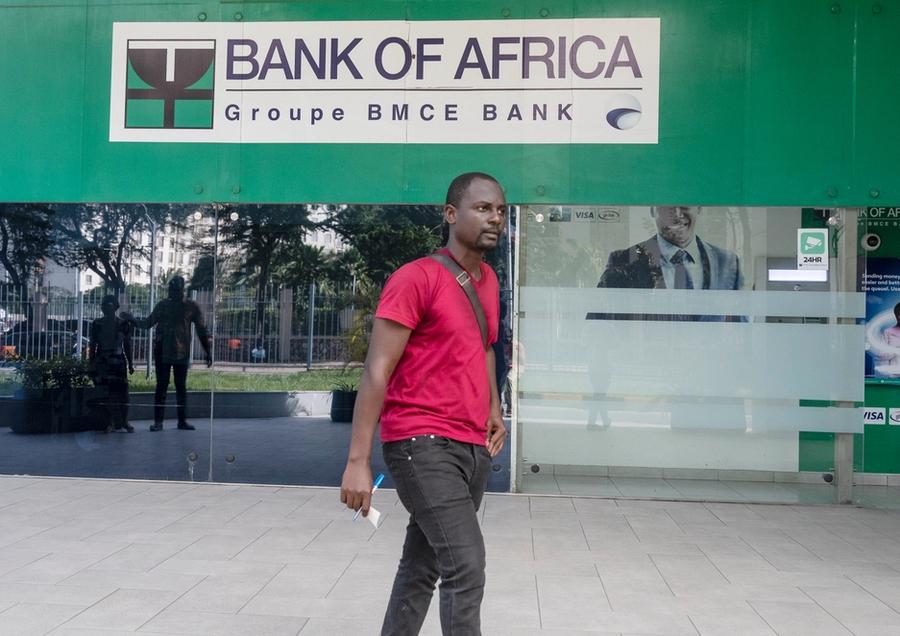 A man walking in front of a Bank of Africa branch in Ghana, wearing a red shirt and holding papers.
