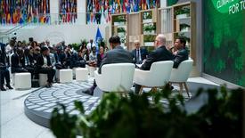 A panel discussion in a conference setting with four speakers seated on stage in front of an audience. The background features a large display with flags from various countries and a green backdrop with text related to climate action.