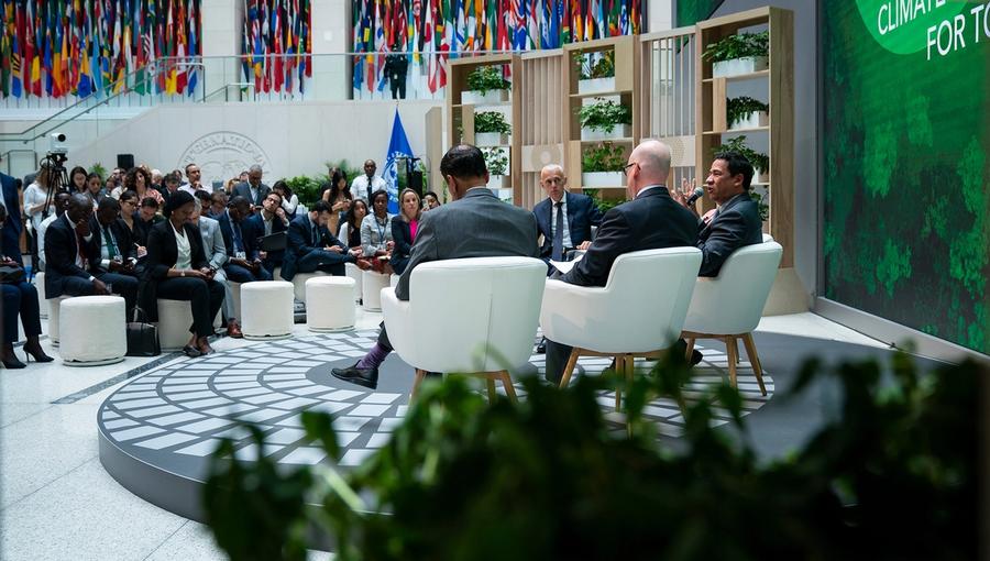 A panel discussion in a conference setting with four speakers seated on stage in front of an audience. The background features a large display with flags from various countries and a green backdrop with text related to climate action.