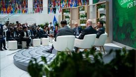 A panel discussion in a conference setting with four speakers seated on stage in front of an audience. The background features a large display with flags from various countries and a green backdrop with text related to climate action.