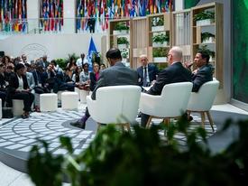 A panel discussion in a conference setting with four speakers seated on stage in front of an audience. The background features a large display with flags from various countries and a green backdrop with text related to climate action.