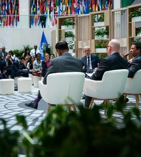 A panel discussion in a conference setting with four speakers seated on stage in front of an audience. The background features a large display with flags from various countries and a green backdrop with text related to climate action.