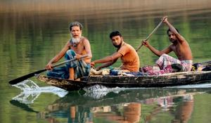 five people rowing a small wooden boat on calm water in Bangladesh, with reflections visible in the water.