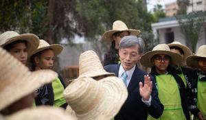 A man in a suit speaking to a group of children wearing green aprons and straw hats in an outdoor setting.