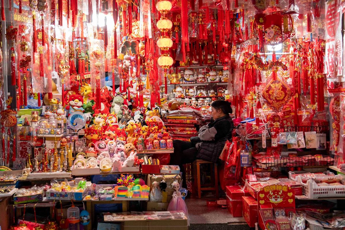A colorful market stall in China filled with various traditional Chinese decorations, lanterns, and toys, predominantly in red and gold, with a person sitting amidst the items.
