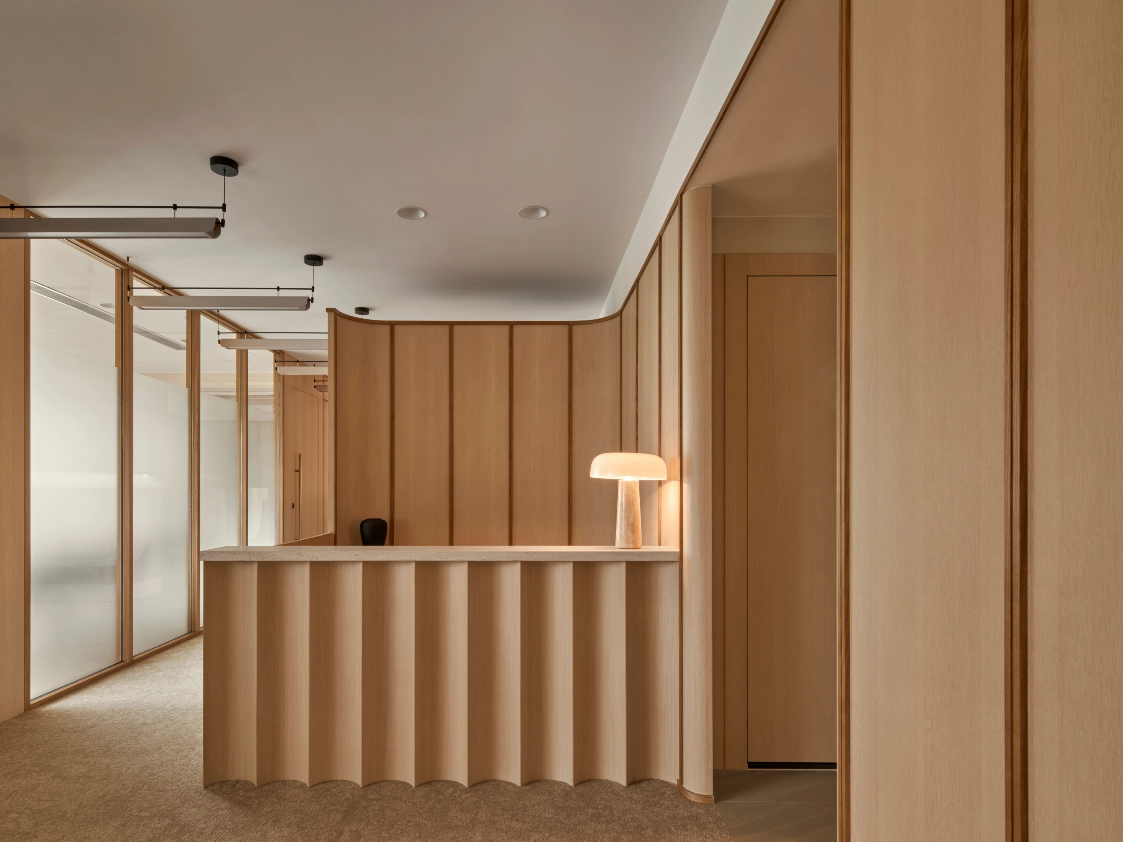 An inviting reception area showcases smooth wooden paneling with gentle curves, a chic marble countertop, and a modern lamp, all accentuated by soft, ambient lighting for a serene ambiance.