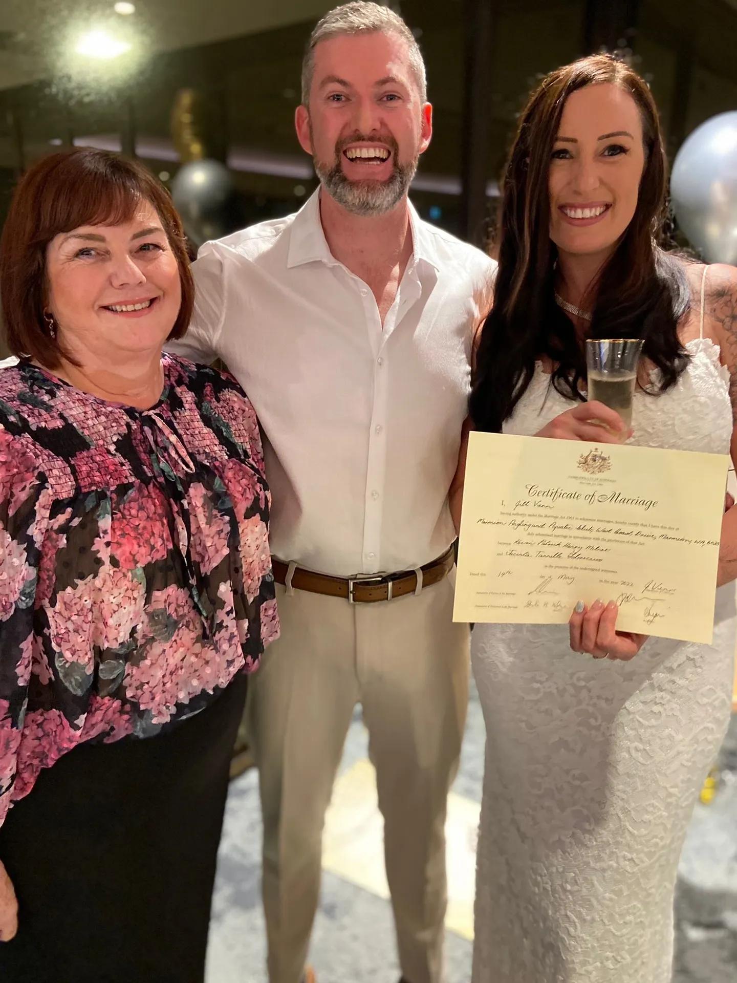 A photo of a man and a woman standing wth their wedding celebrant, holding their celebratory wedding certificate.