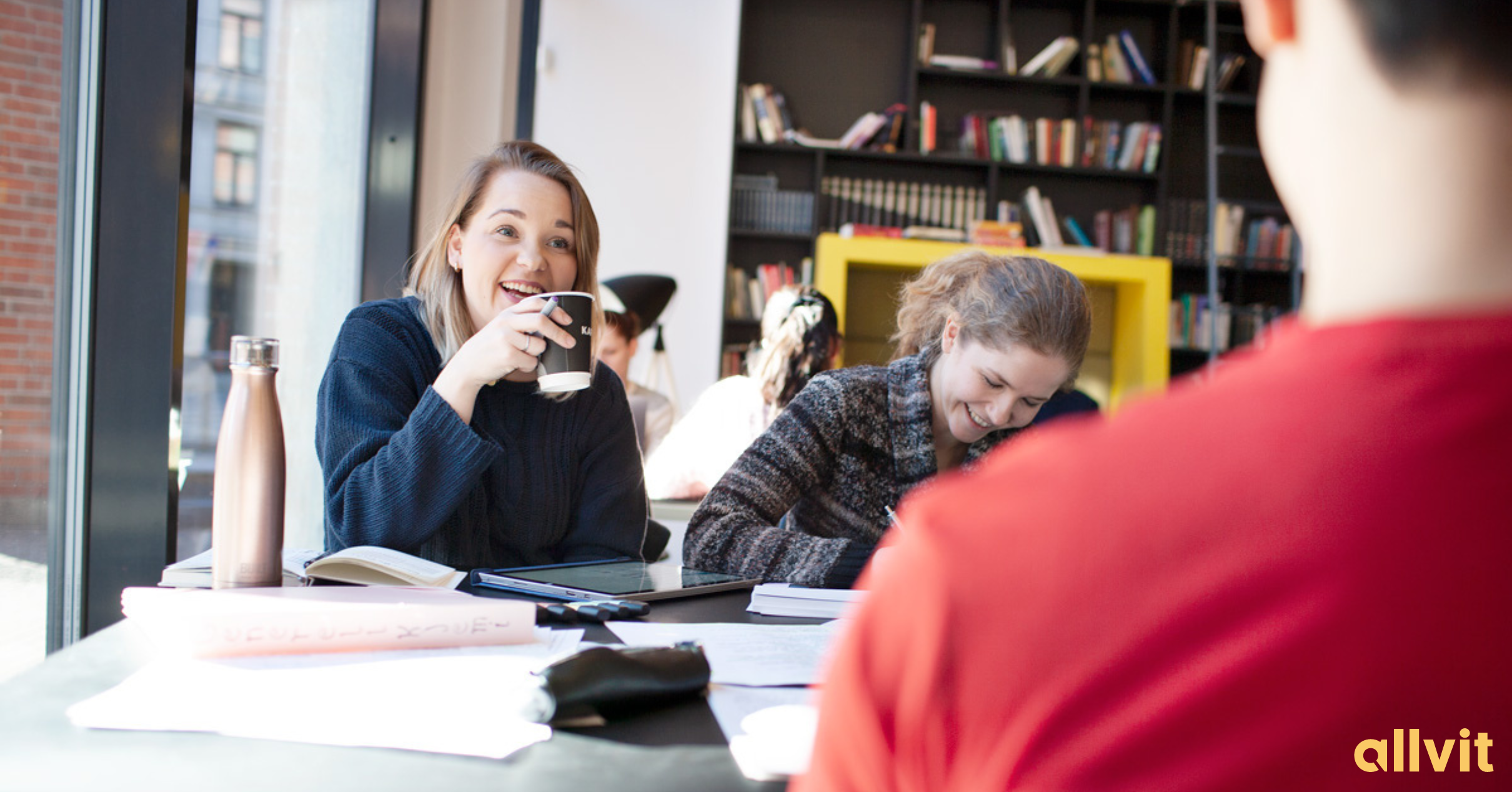 To jenter som smiler til gutt som sitter med ryggen mot kamera på biblioteket. Foto.