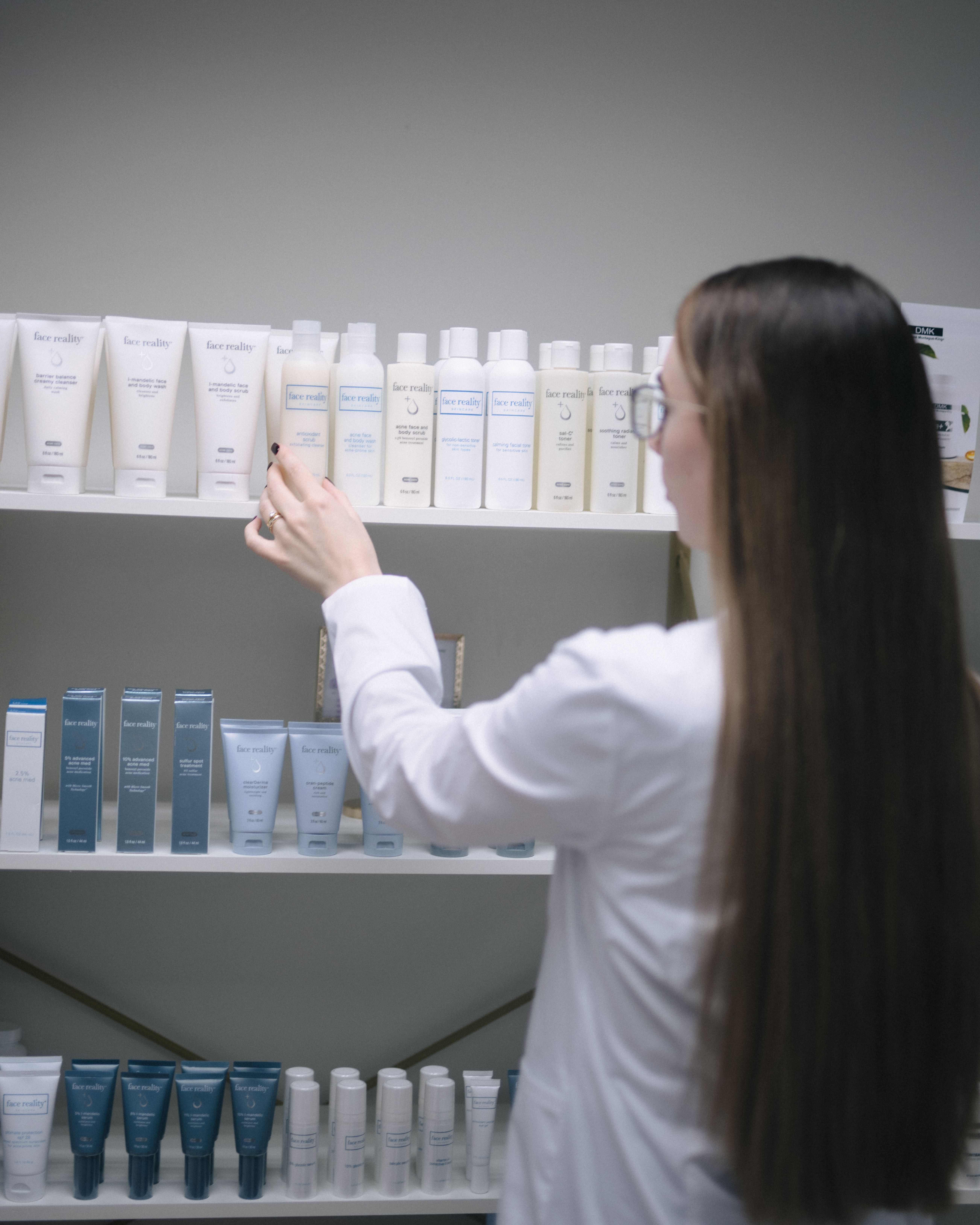 A person in a white lab coat reaching for a bottle of Face Reality skincare product on a shelf.