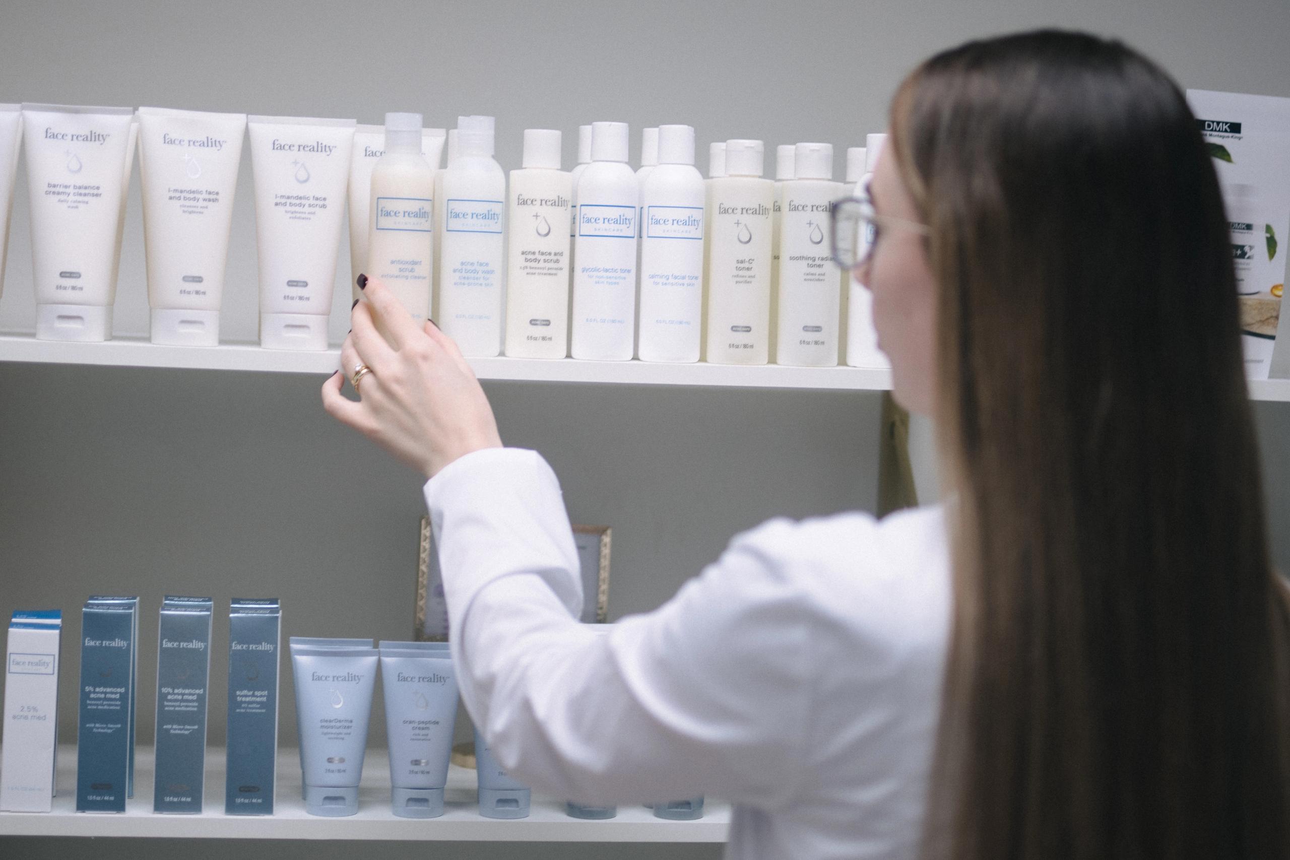 A person in a white lab coat reaching for a bottle of Face Reality skincare product on a shelf.