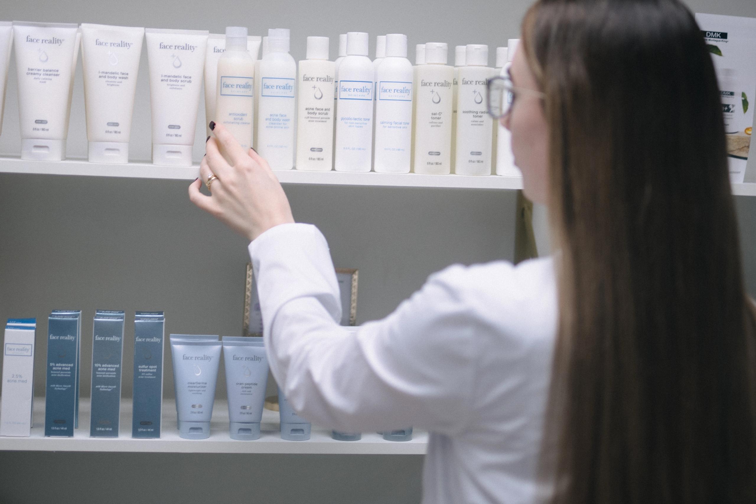 An esthetician reaches for a skincare product on a shelf filled with "Face Reality" brand items.