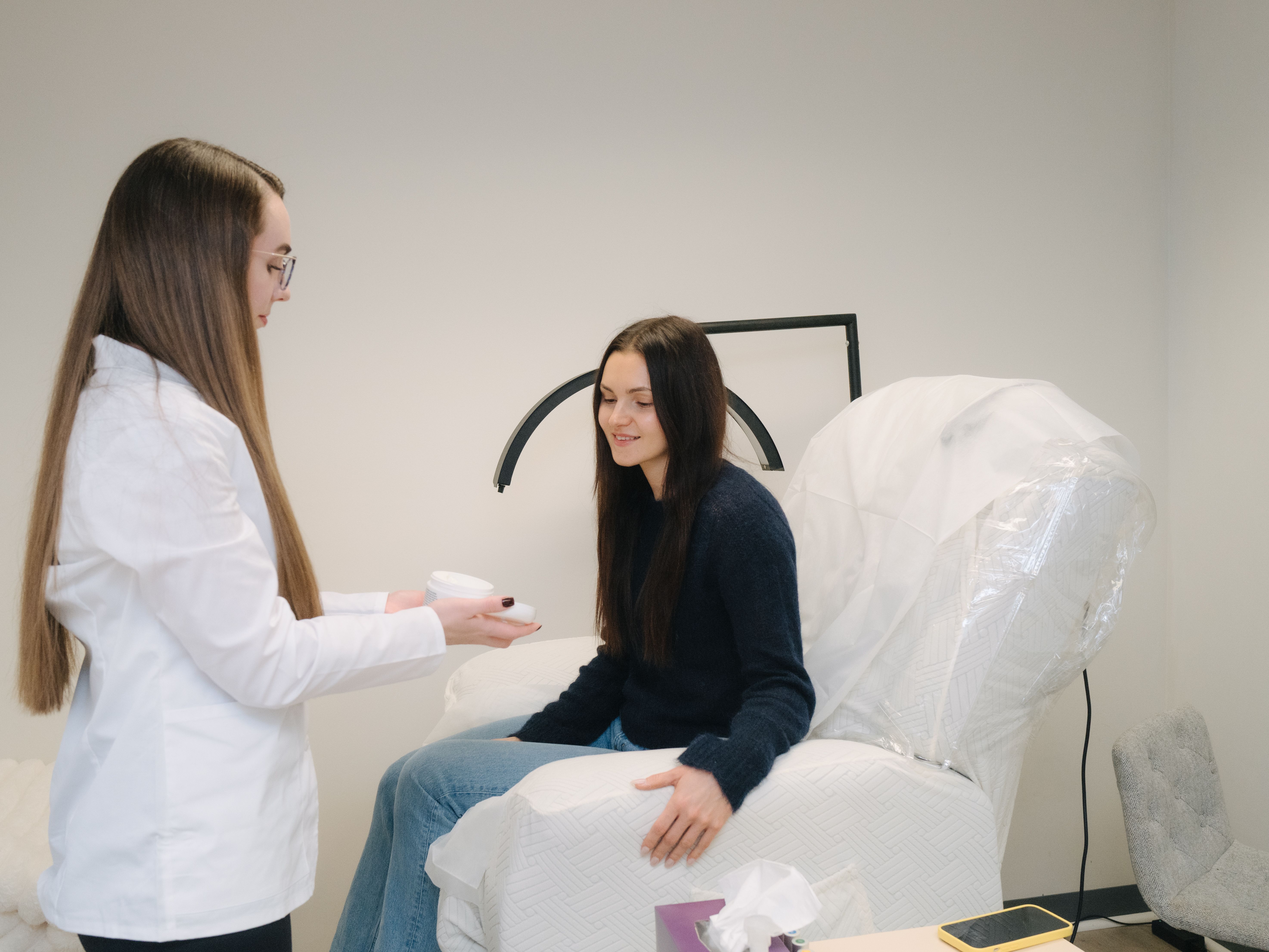 A professional in a white coat presents a container to a smiling client seated on a treatment bed.