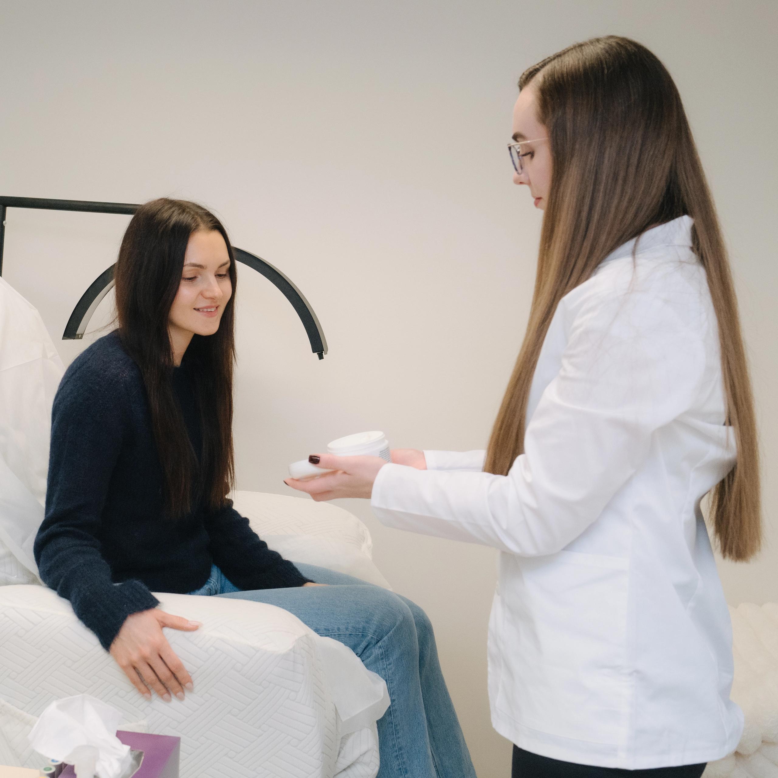 A professional in a white coat presents a container to a smiling client seated on a treatment bed.