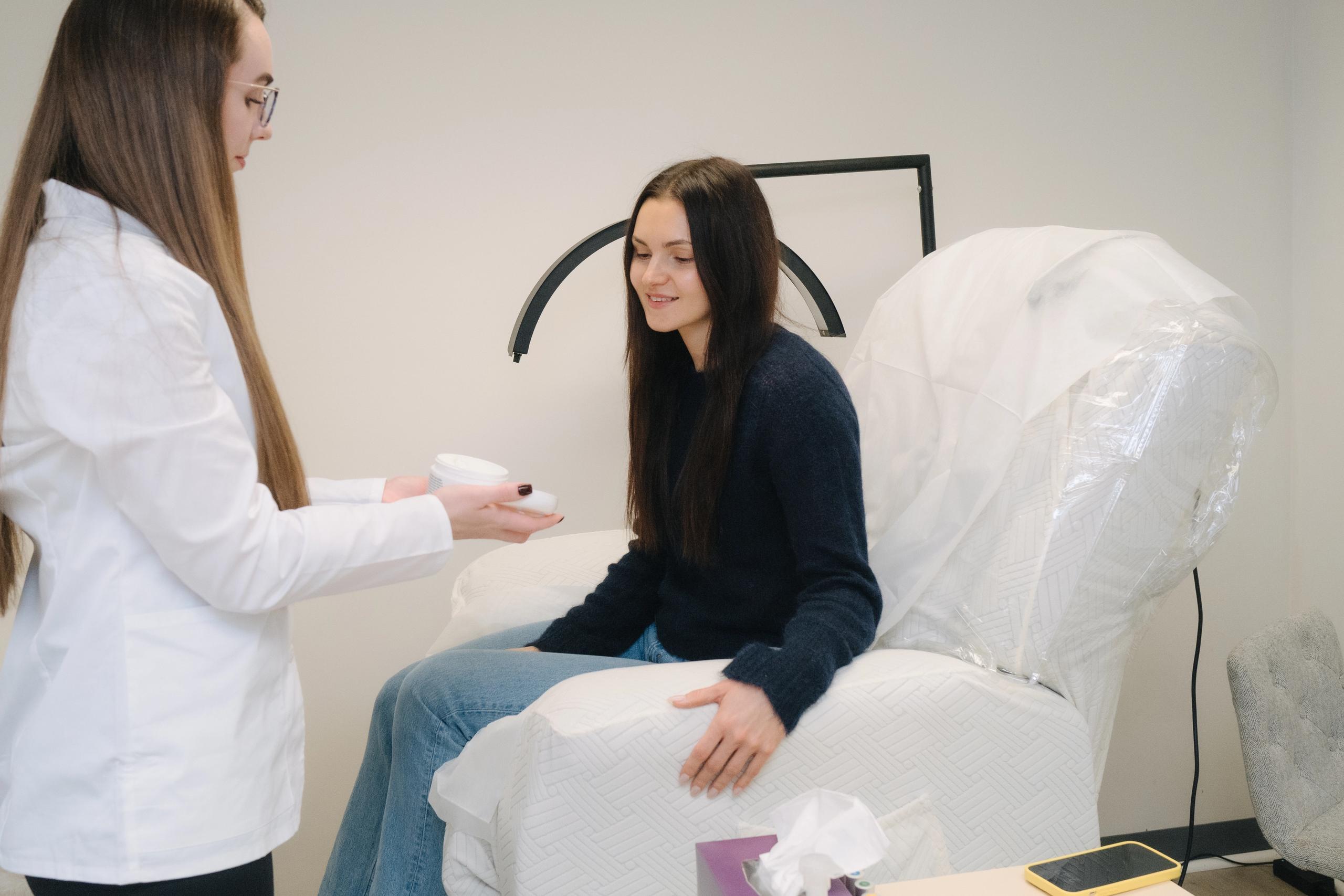 An esthetician presents a white container to a smiling woman seated on a recliner chair.