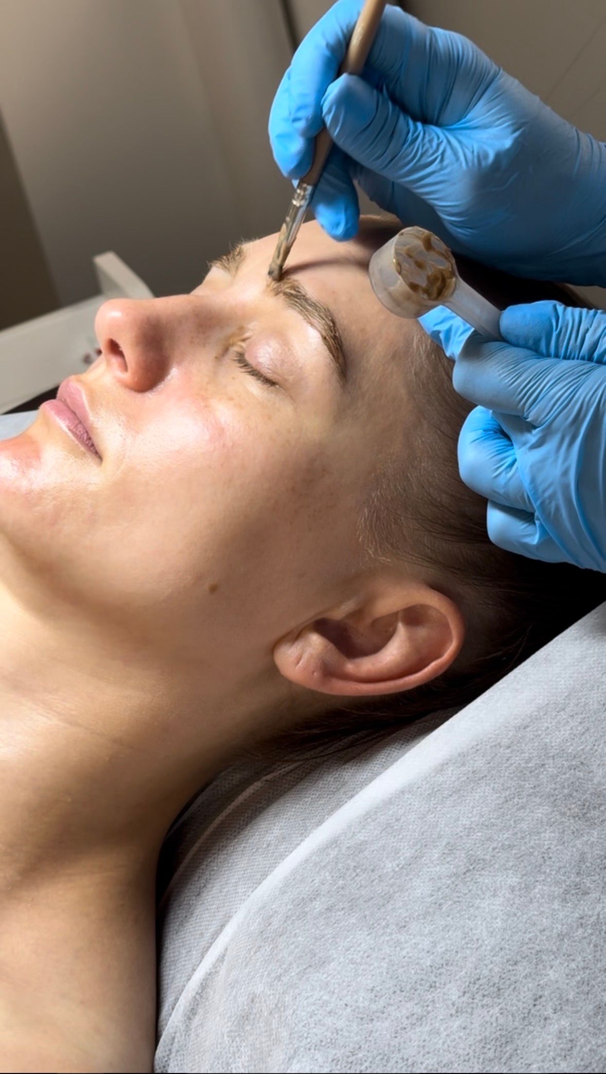 A person lies with eyes closed as gloved hands apply a brow tint with a small brush.