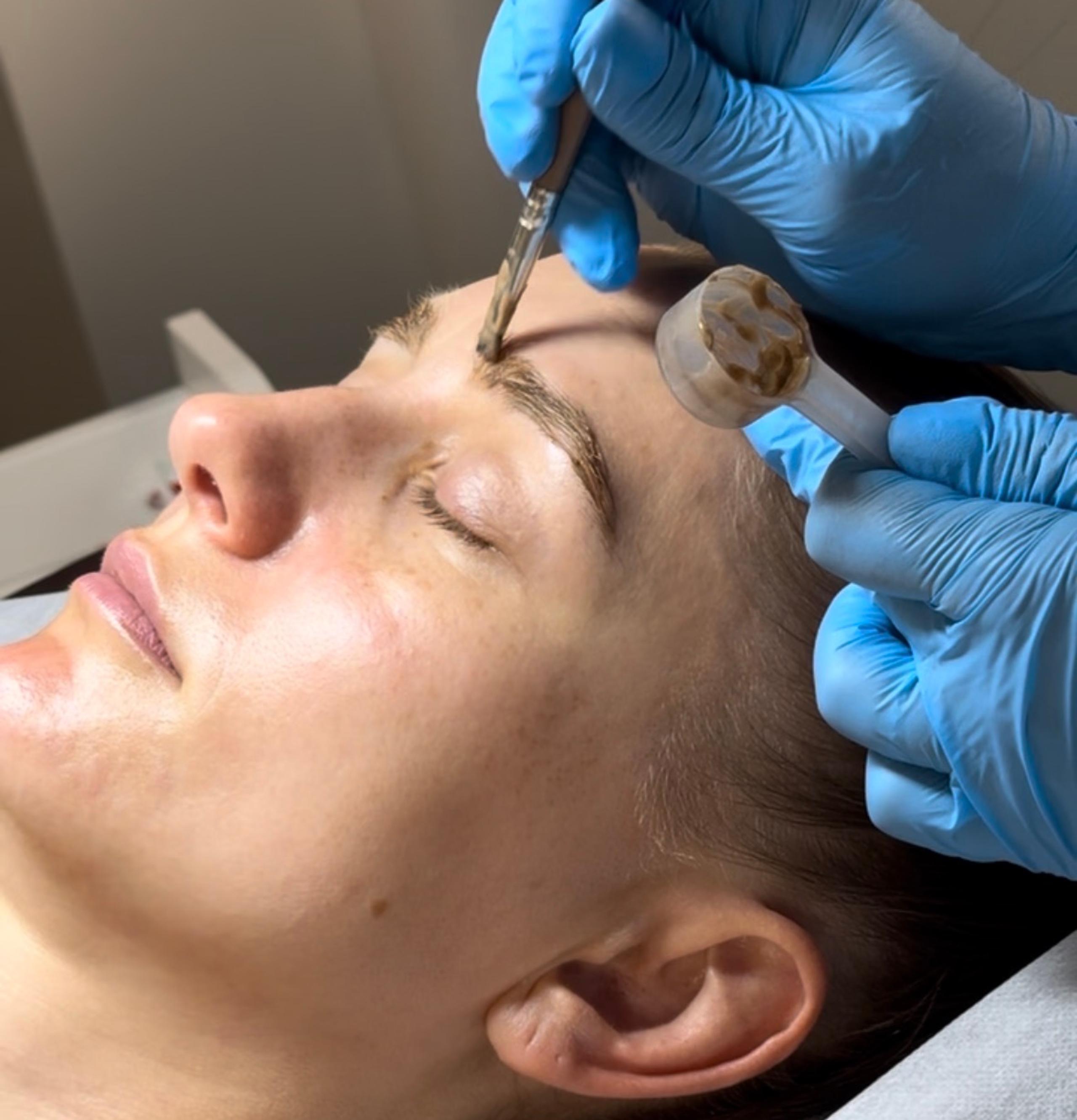 A person lies with eyes closed as gloved hands apply a brow tint with a small brush.