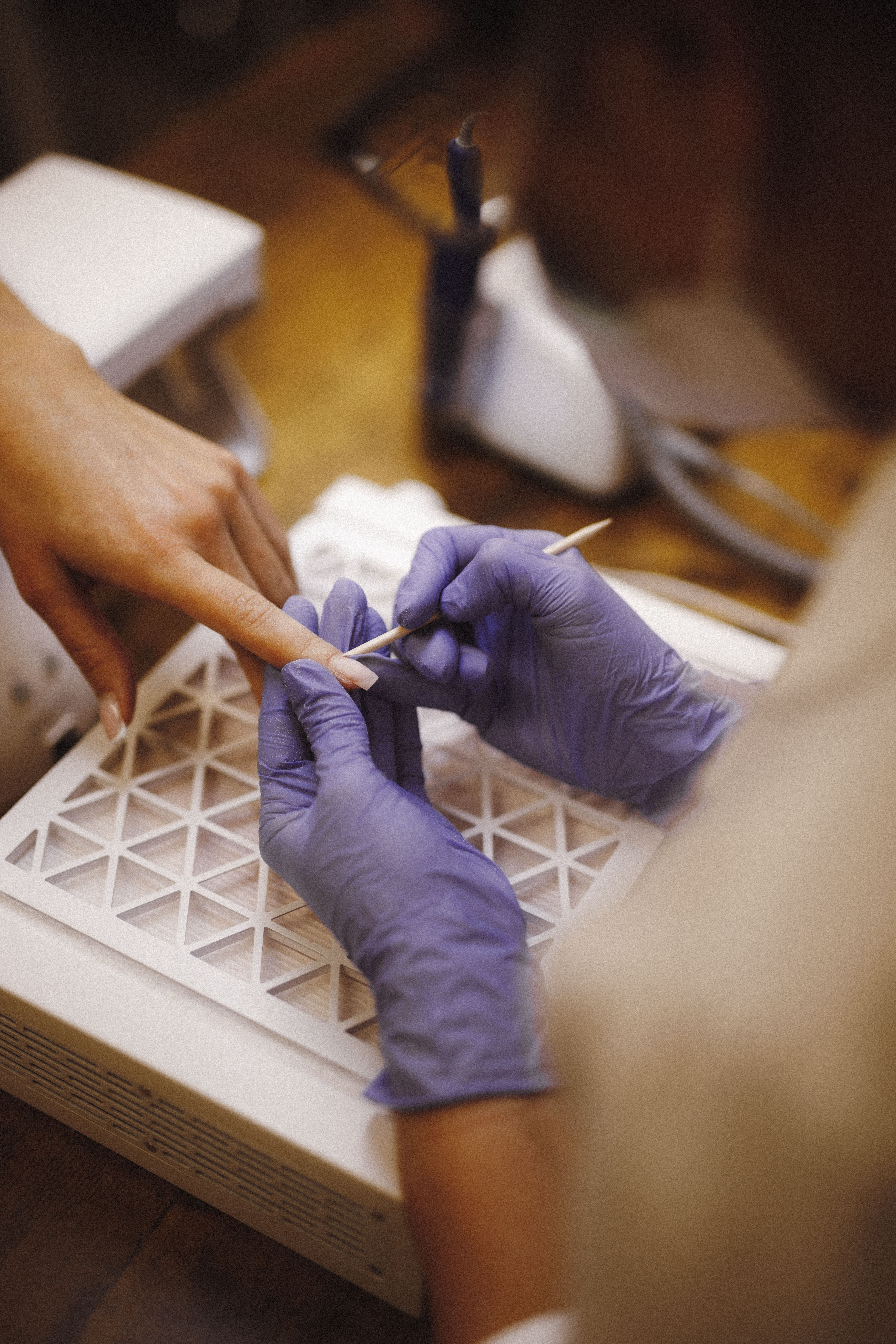 A nail technician in purple gloves uses a wooden stick to push back a client's cuticles.