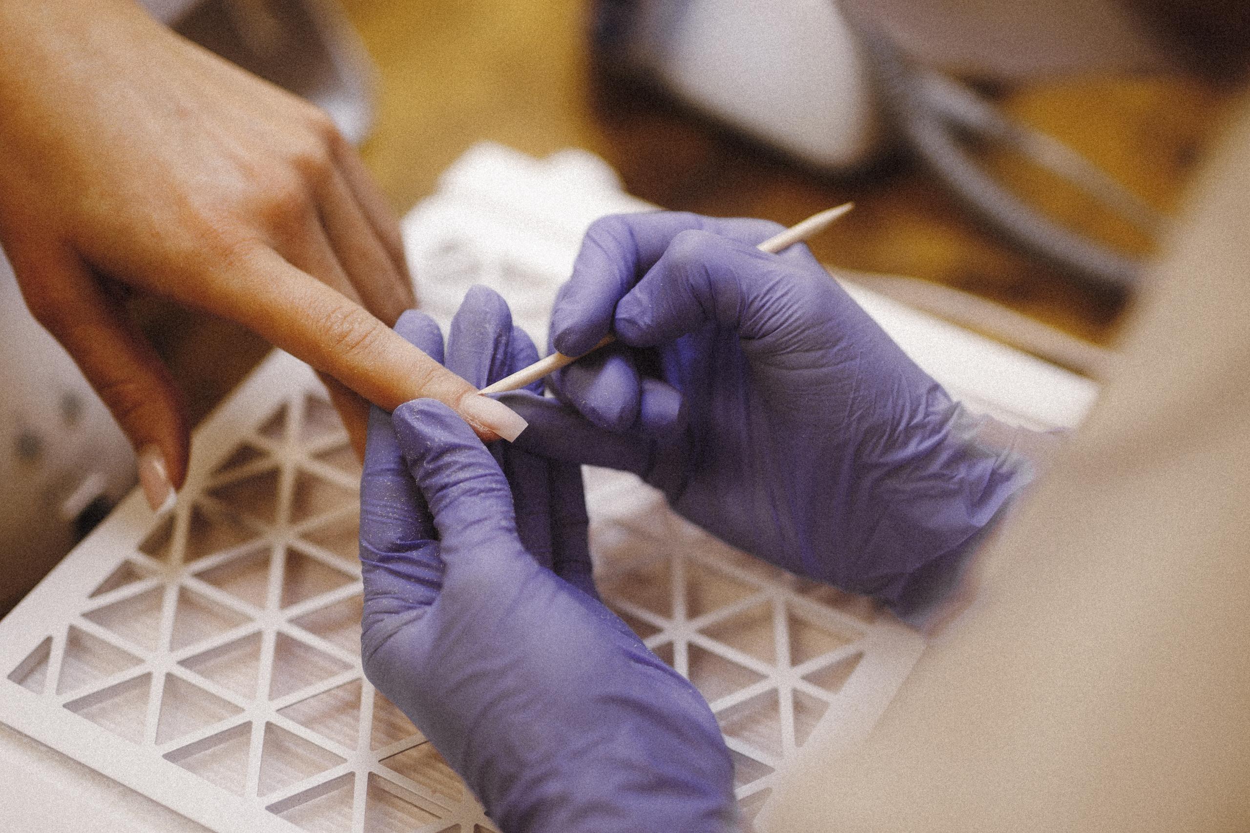 A nail technician in purple gloves uses a wooden stick to push back a client's cuticles.