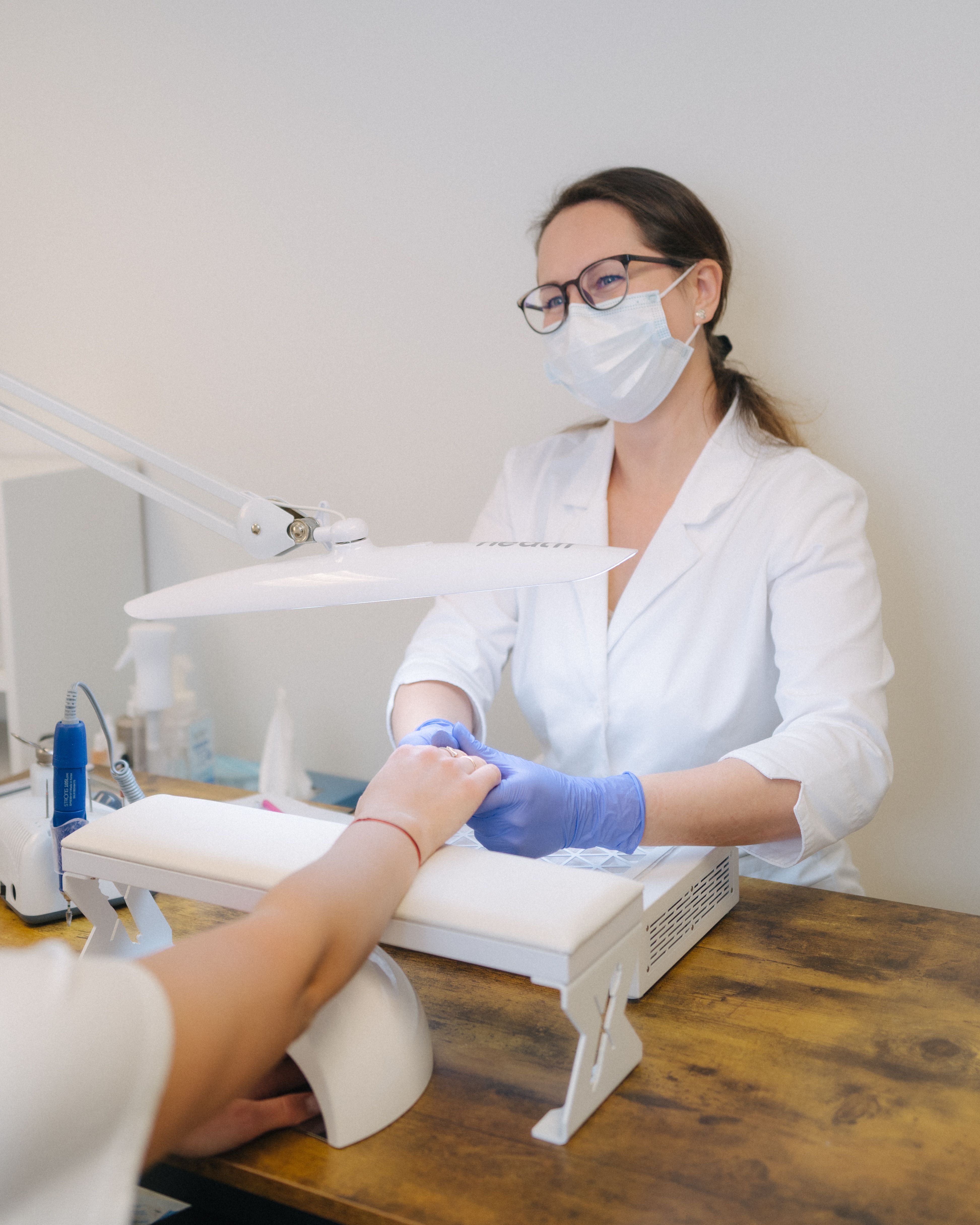 A masked nail technician in gloves works on a customer's hand at a nail salon station.