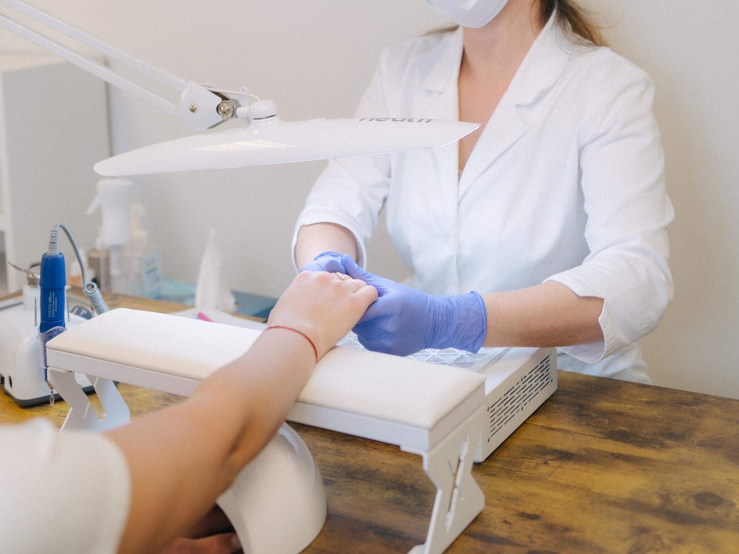 A masked nail technician in gloves works on a customer's hand at a nail salon station.