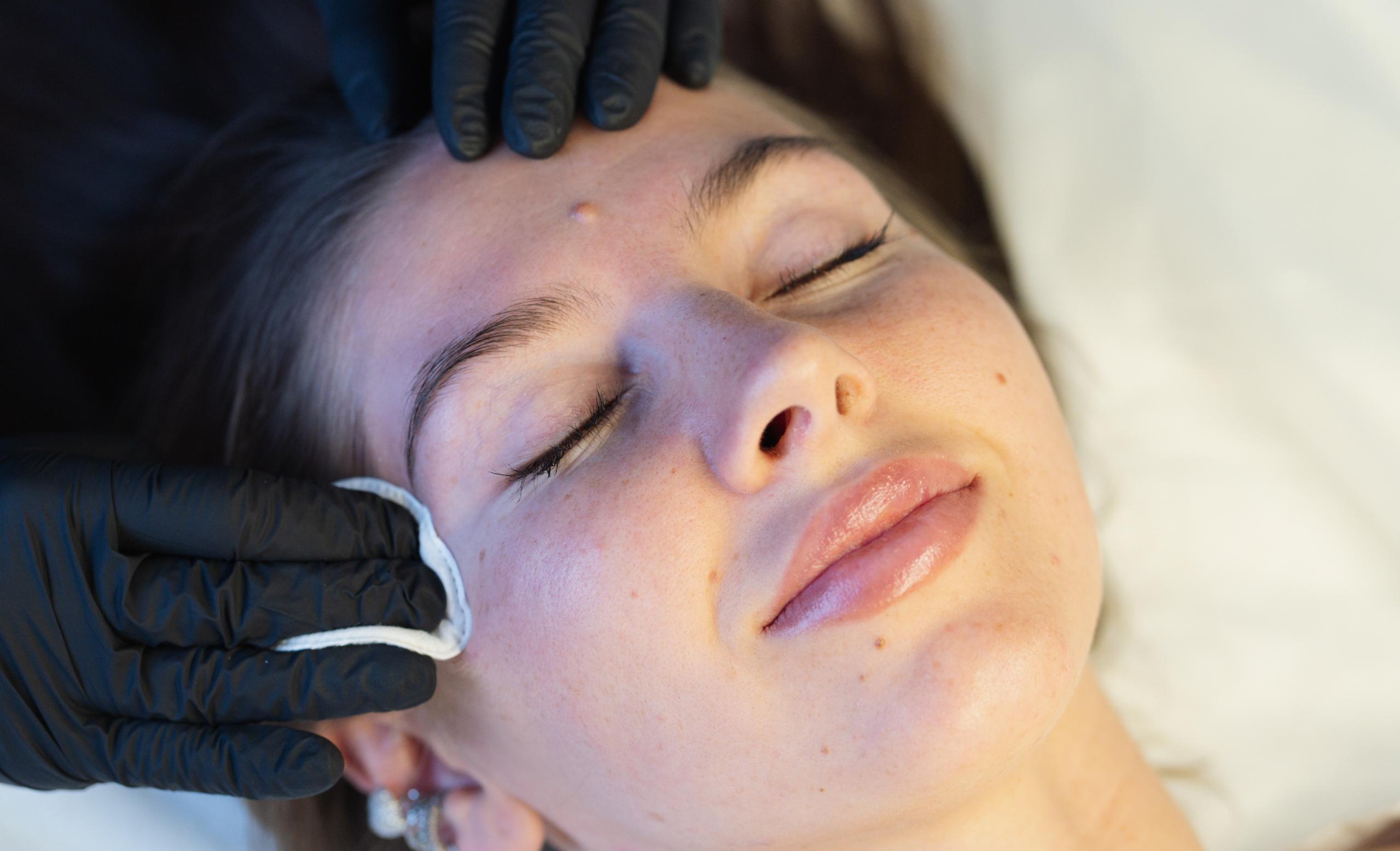 An esthetician wipes an eyebrow of a client, who is laying on the treatment bed