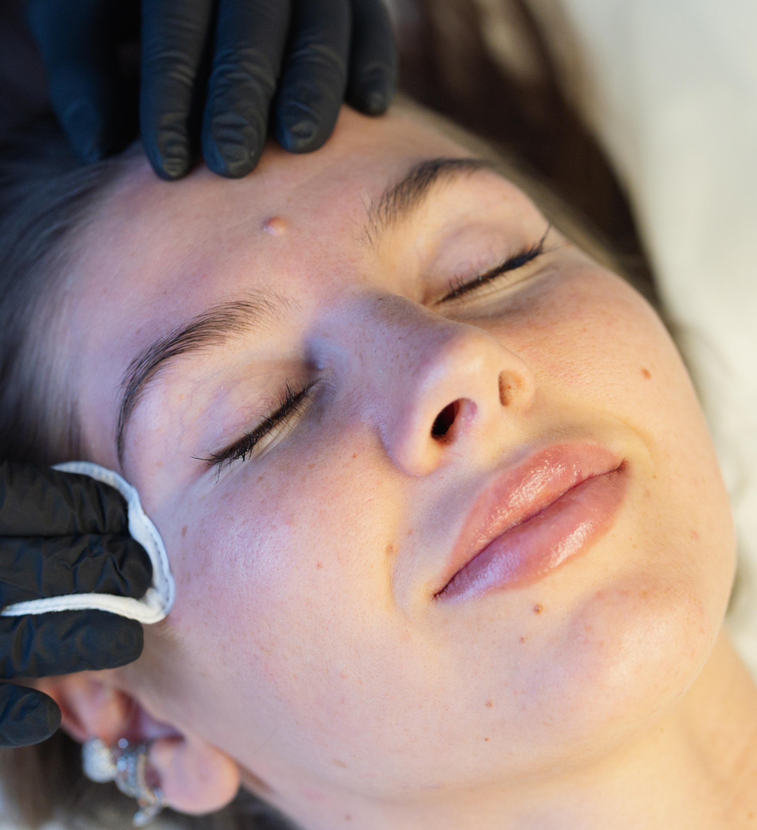 Esthetician in black gloves cleans a client's eyebrow with a cotton pad before brow tinting.