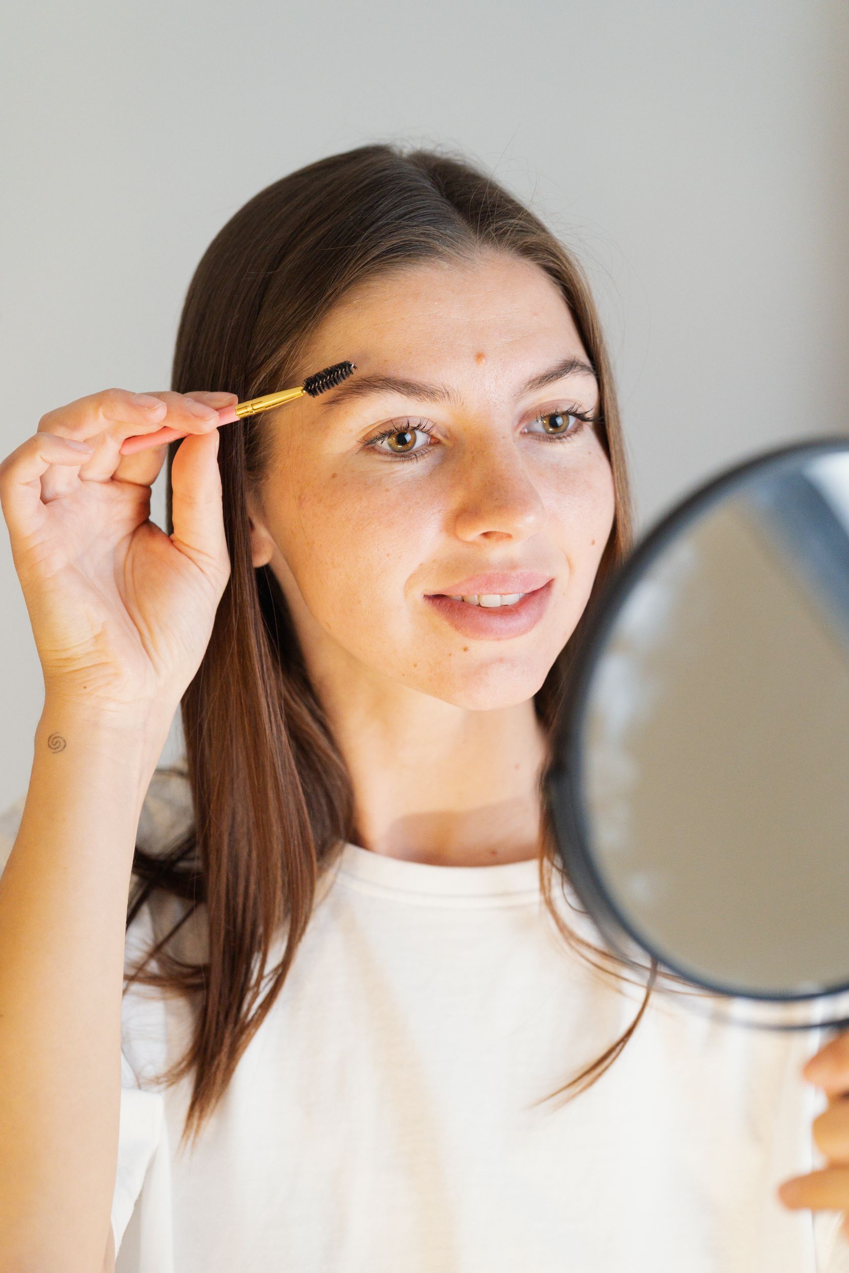 A woman brushes her eyebrow with a spoolie while looking in a mirror.