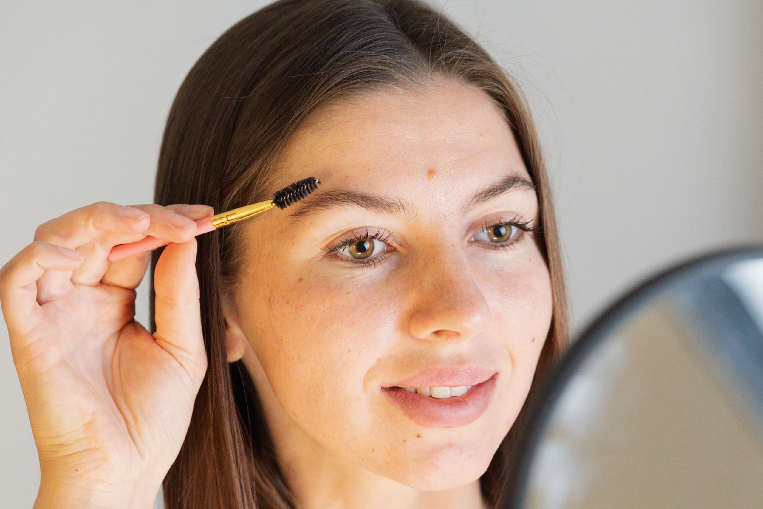 A woman brushes her eyebrow with a spoolie while looking in a mirror.