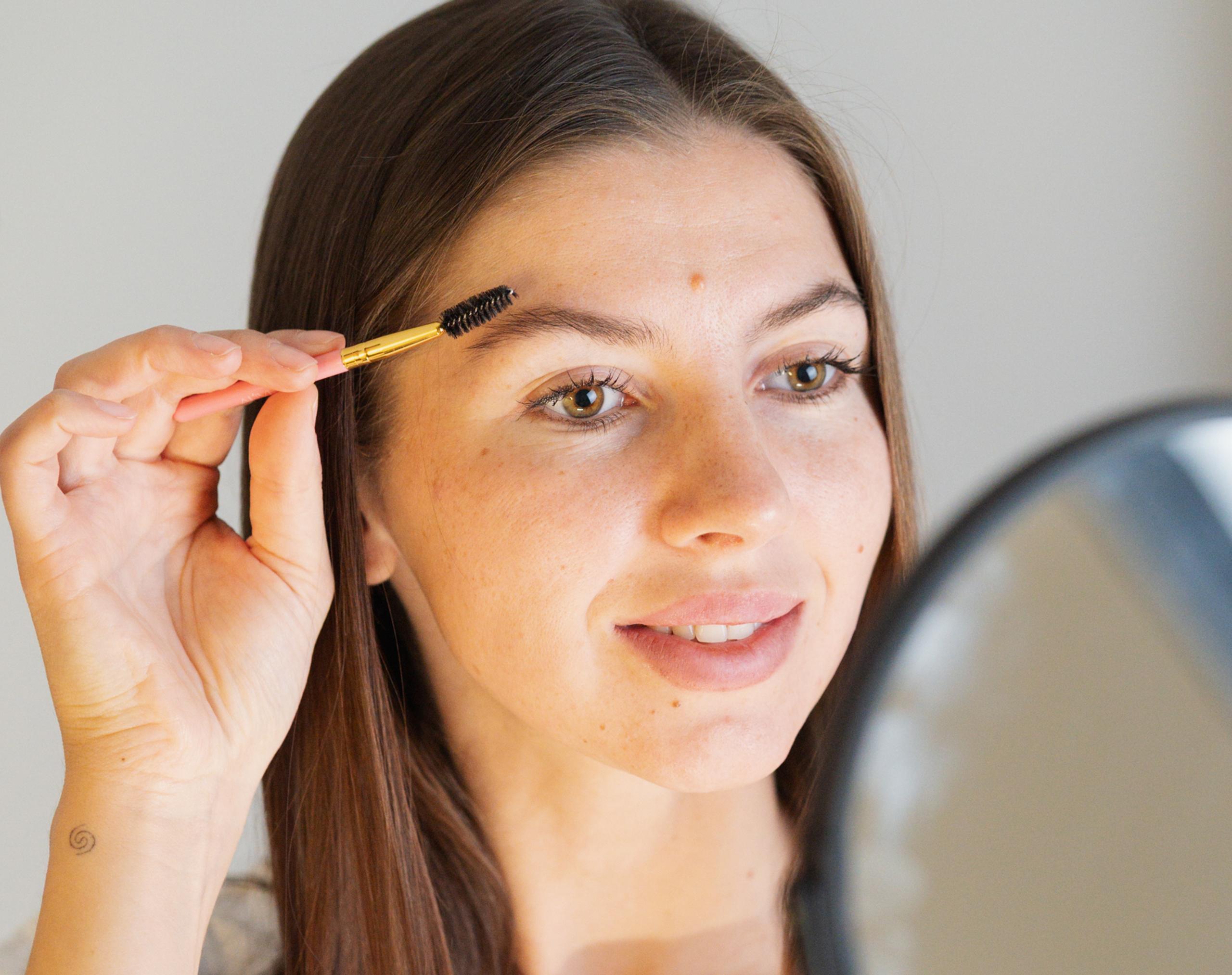 A woman brushes her eyebrow with a spoolie while looking in a mirror.