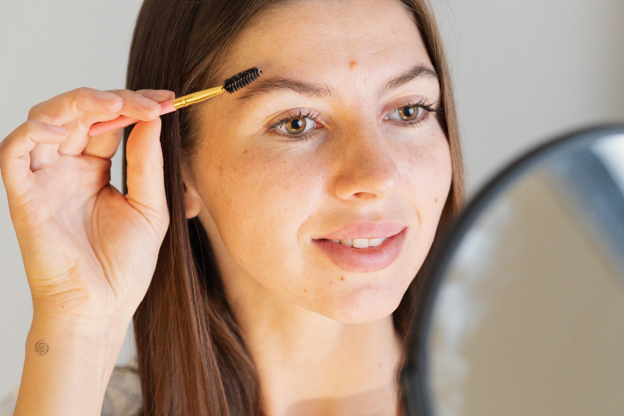 A woman brushes her eyebrow with a spoolie while looking in a mirror.