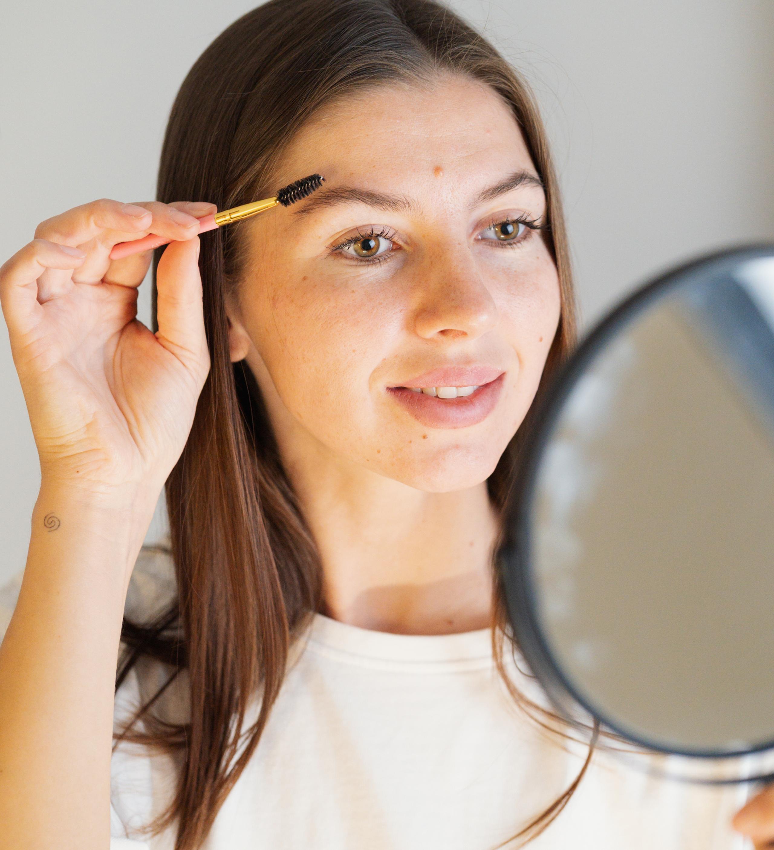 A woman looking in a mirror while brushing her eyebrow with a spoolie brush