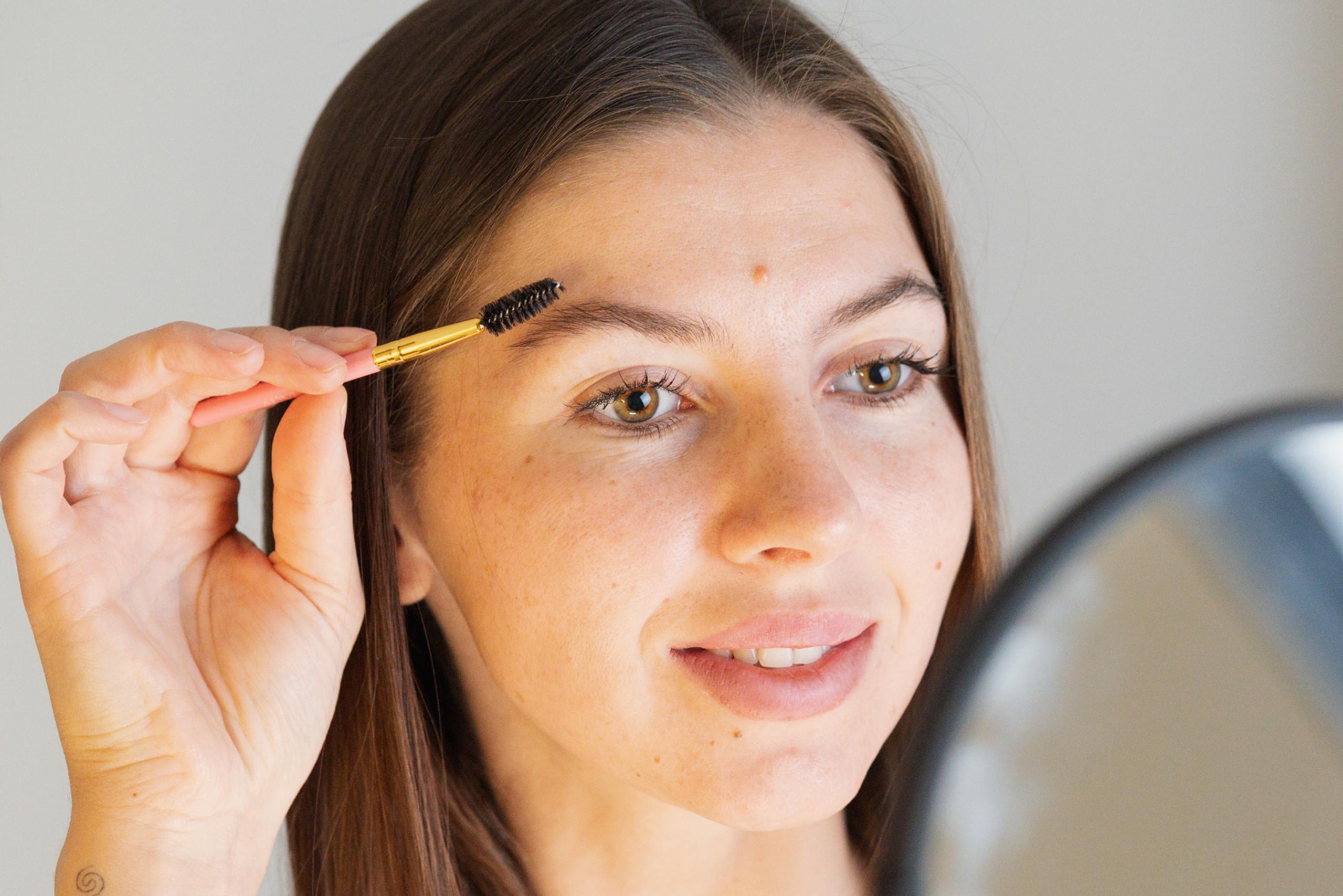 A woman uses a spoolie brush to groom her eyebrow while looking in a mirror.