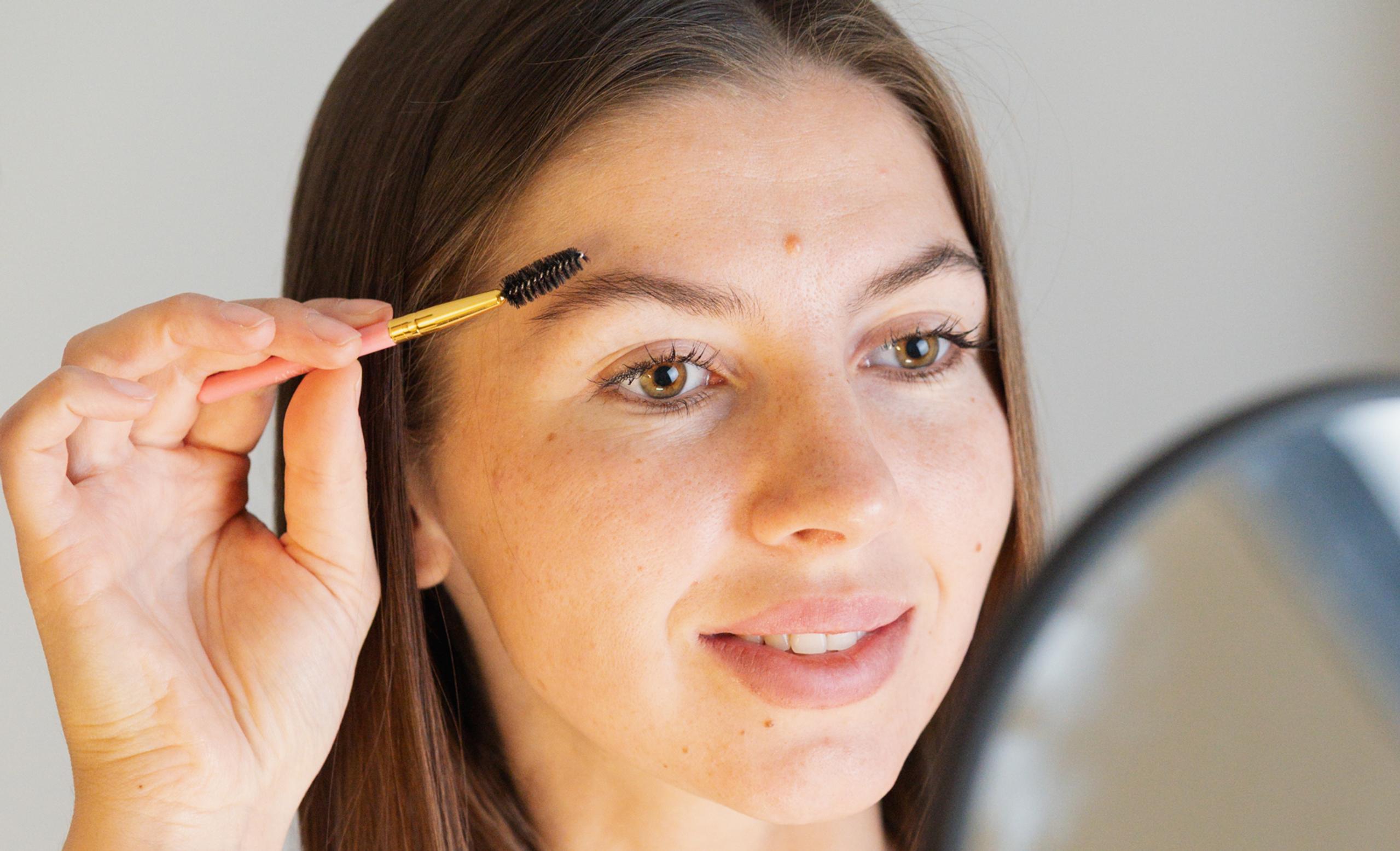 A woman uses a spoolie brush to groom her eyebrow while looking in a mirror.