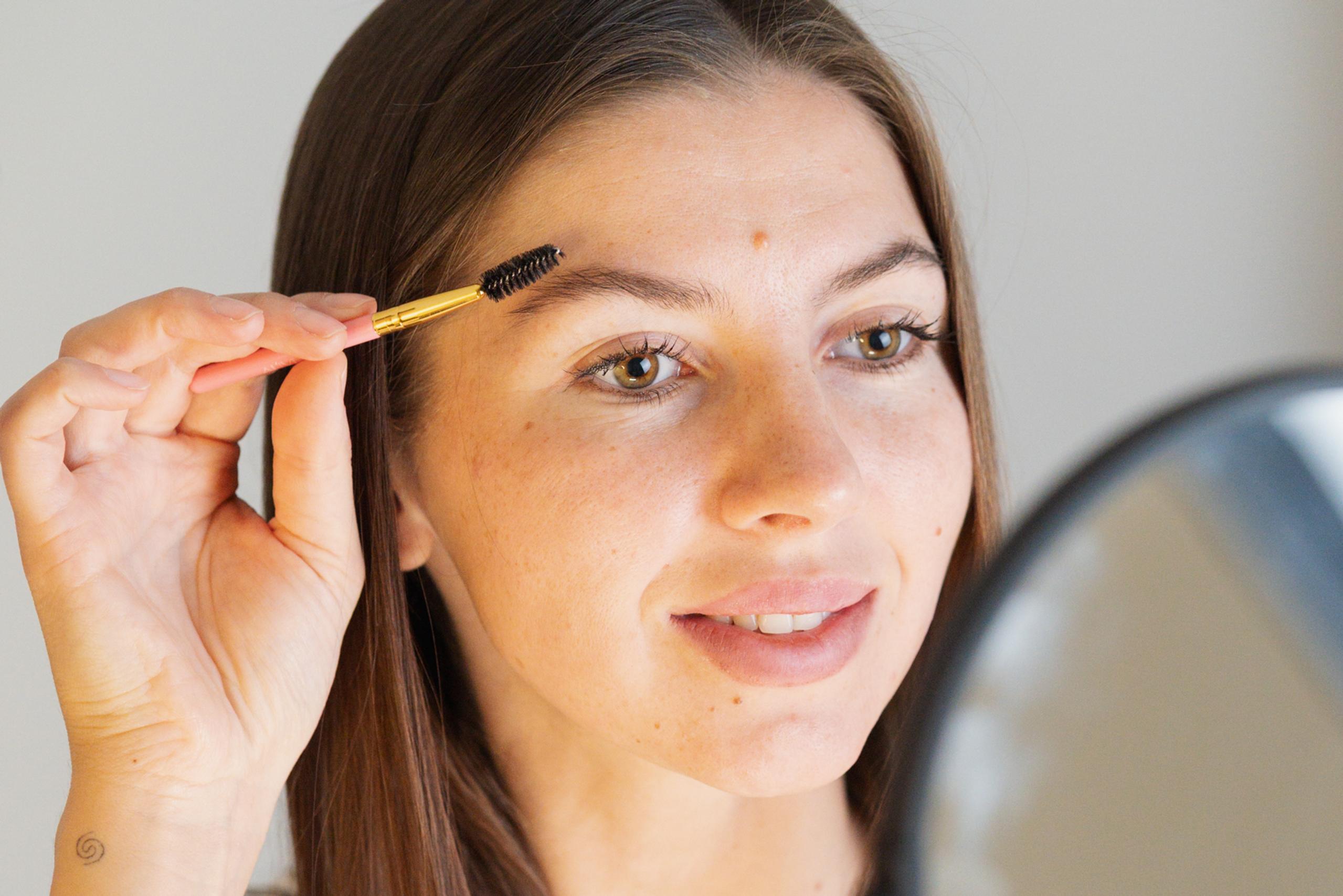 Client brushes her eyebrows with a spoolie while looking in a mirror before her appointment.