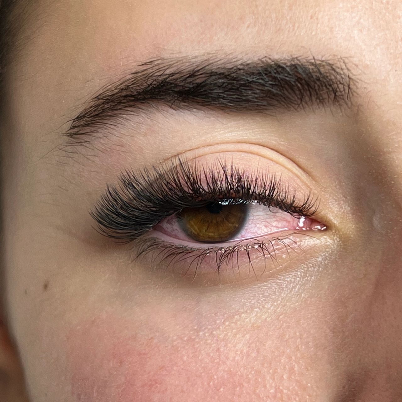 Close-up of a brown eye with thick, dark eyelash extensions and a full eyebrow; the eye appears red.