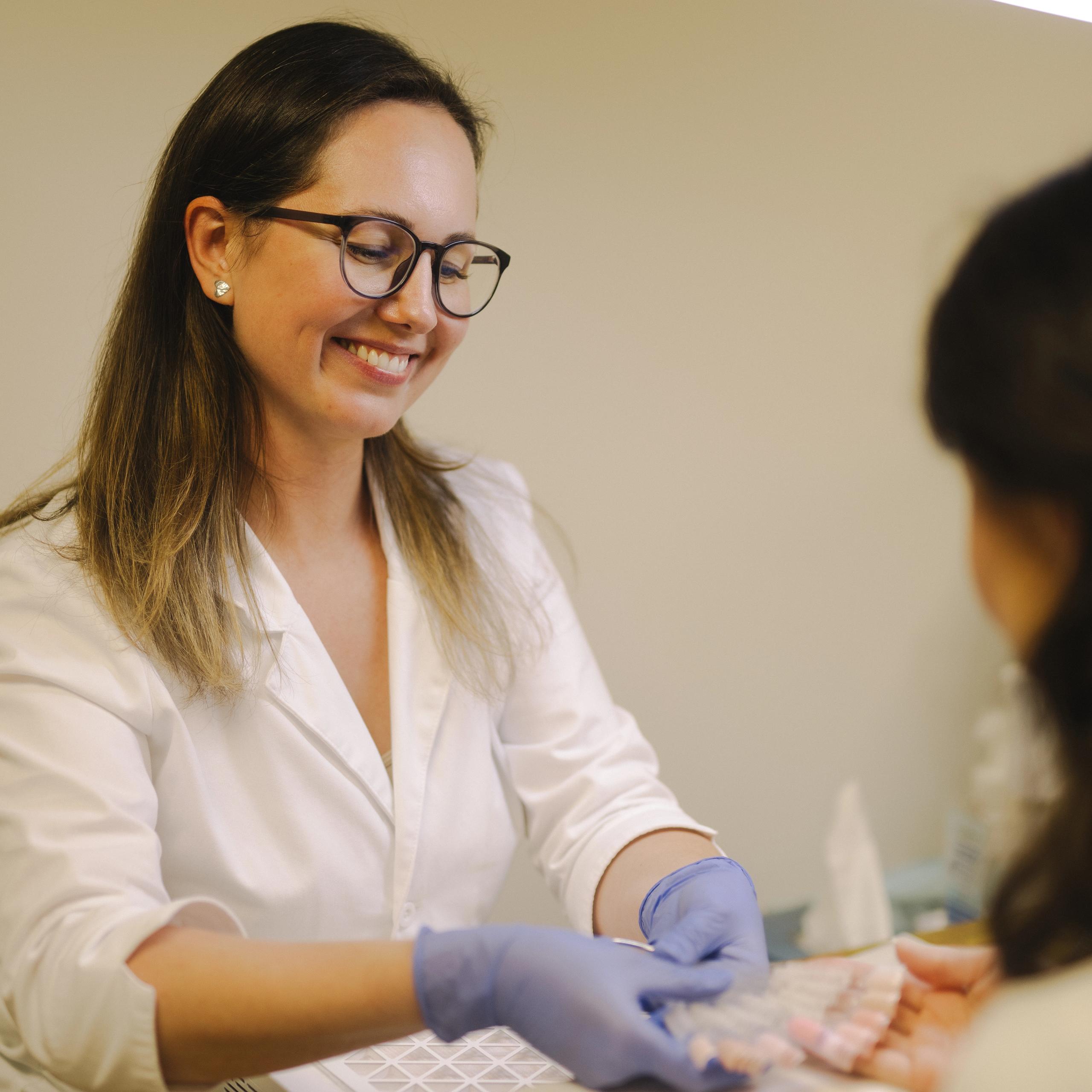 A smiling nail technician in blue gloves presents nail polish samples to a client.